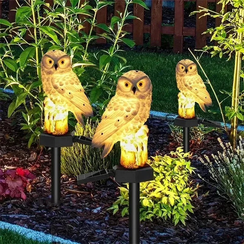 Three owl lights glowing warm yellow on stakes among shrubs and bark chippings in a garden at night. 7