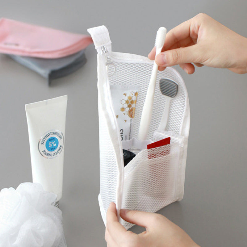 Hands zipping open a white mesh pouch standing upright, holding a toothbrush, toothpaste and small items beside a cleanser tube and bath puff. 1