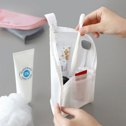 Hands zipping open a white mesh pouch standing upright, holding a toothbrush, toothpaste and small items beside a cleanser tube and bath puff. 1