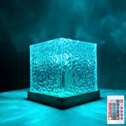 Rippled glass cube lamp with a wooden base glowing turquoise in a dark room, with its small remote control beside it. 1