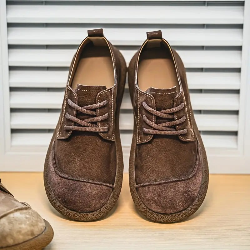 Front view of a pair of brown suede lace-up shoes with contrast stitching, standing on a wooden shelf in front of slatted shutters. 10