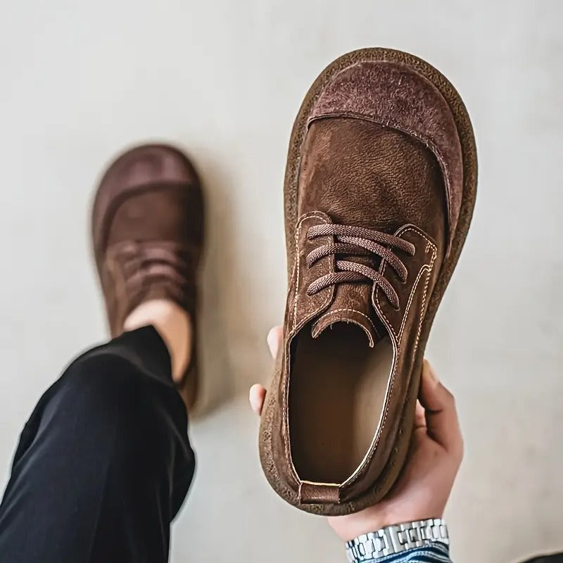 A hand holds a brown shoe tilted towards the camera to show the soft lining and stitching, with the wearer's other foot behind. 7