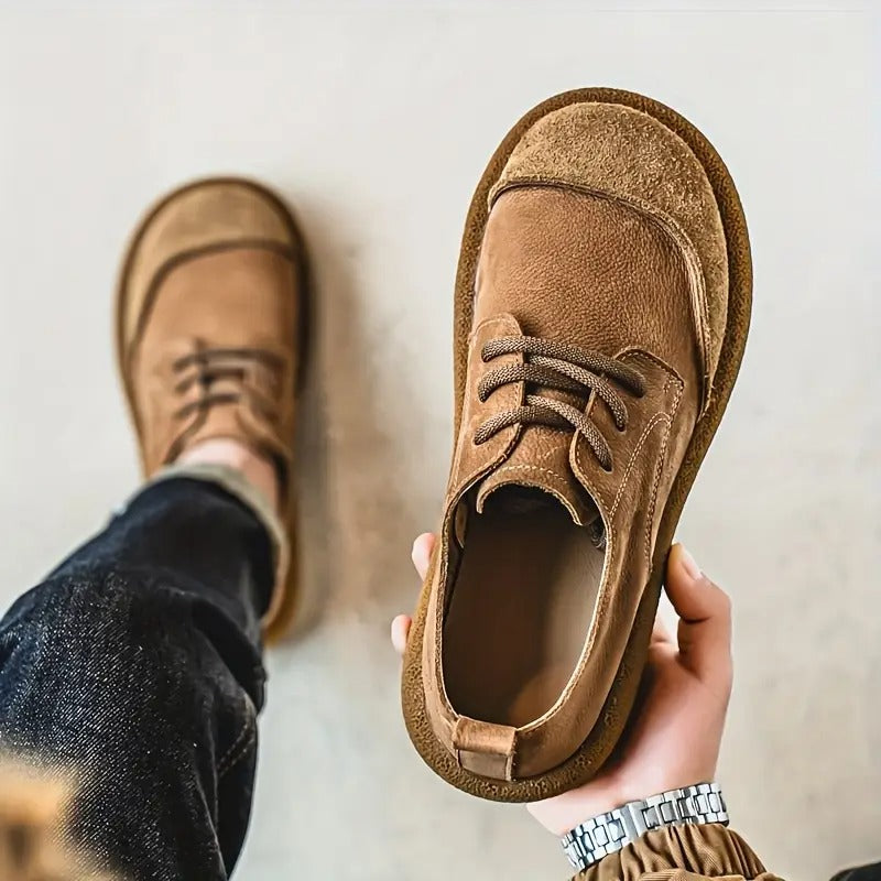 A hand holds a tan shoe angled to show its suede toe cap and stitching, with a wearer's other foot behind. 5