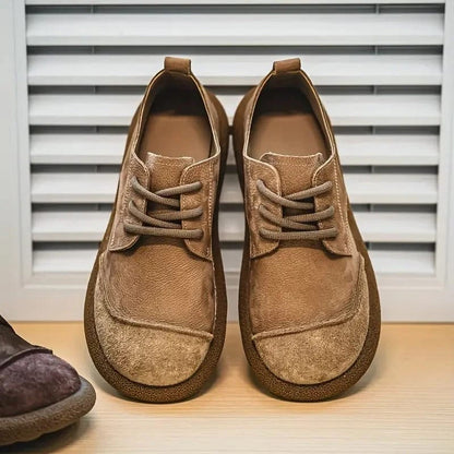Pair of tan suede lace-up shoes from the front on a wooden shelf, with a darker shoe at the edge. 7