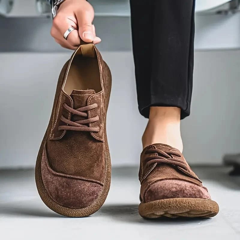 Dark brown suede lace-up shoe lifted by the heel tab beside a worn shoe, with black trousers and a pale background. 8
