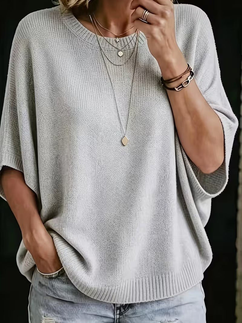 Close-up front view of the light grey batwing knit top worn with layered necklaces, one hand raised to the chin. 1