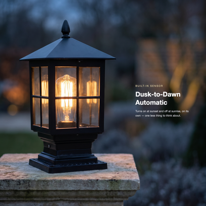 Lit black pillar lamp at dusk on a weathered stone post, with a garden blurred behind and a dusk-to-dawn sensor caption. 2