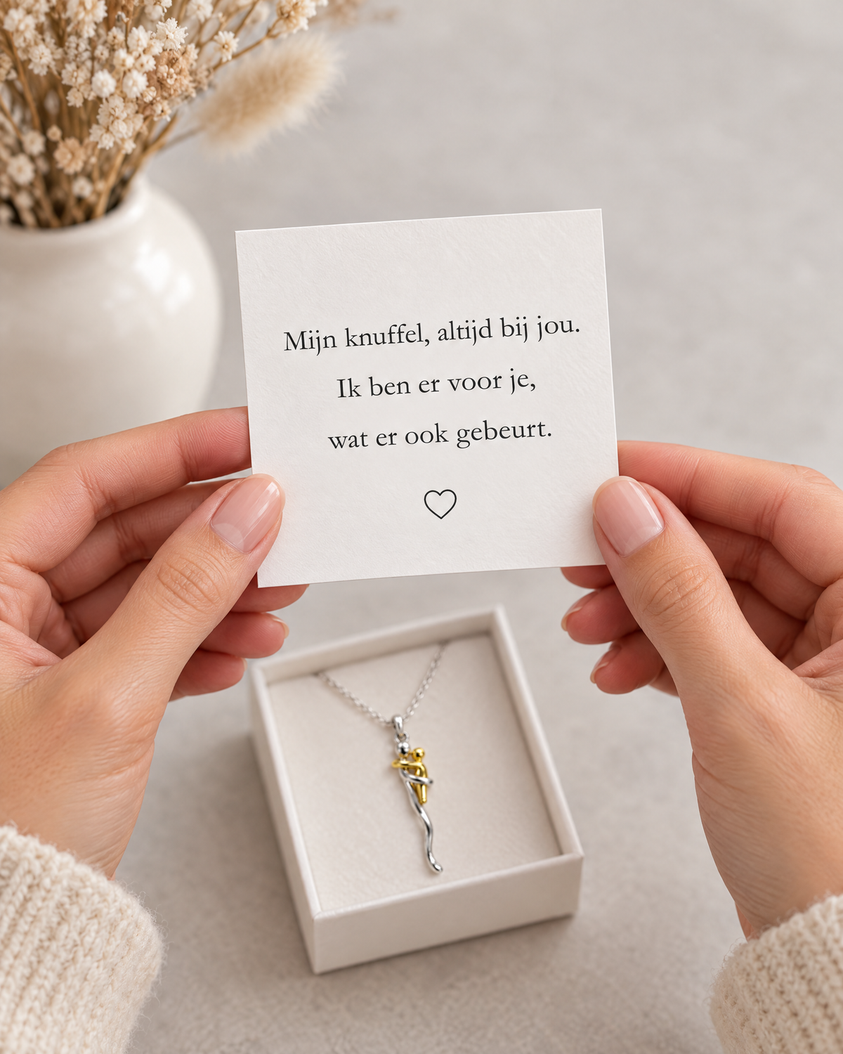 Hands holding a printed message card above an open white gift box that holds the necklace, with dried flowers behind. 6