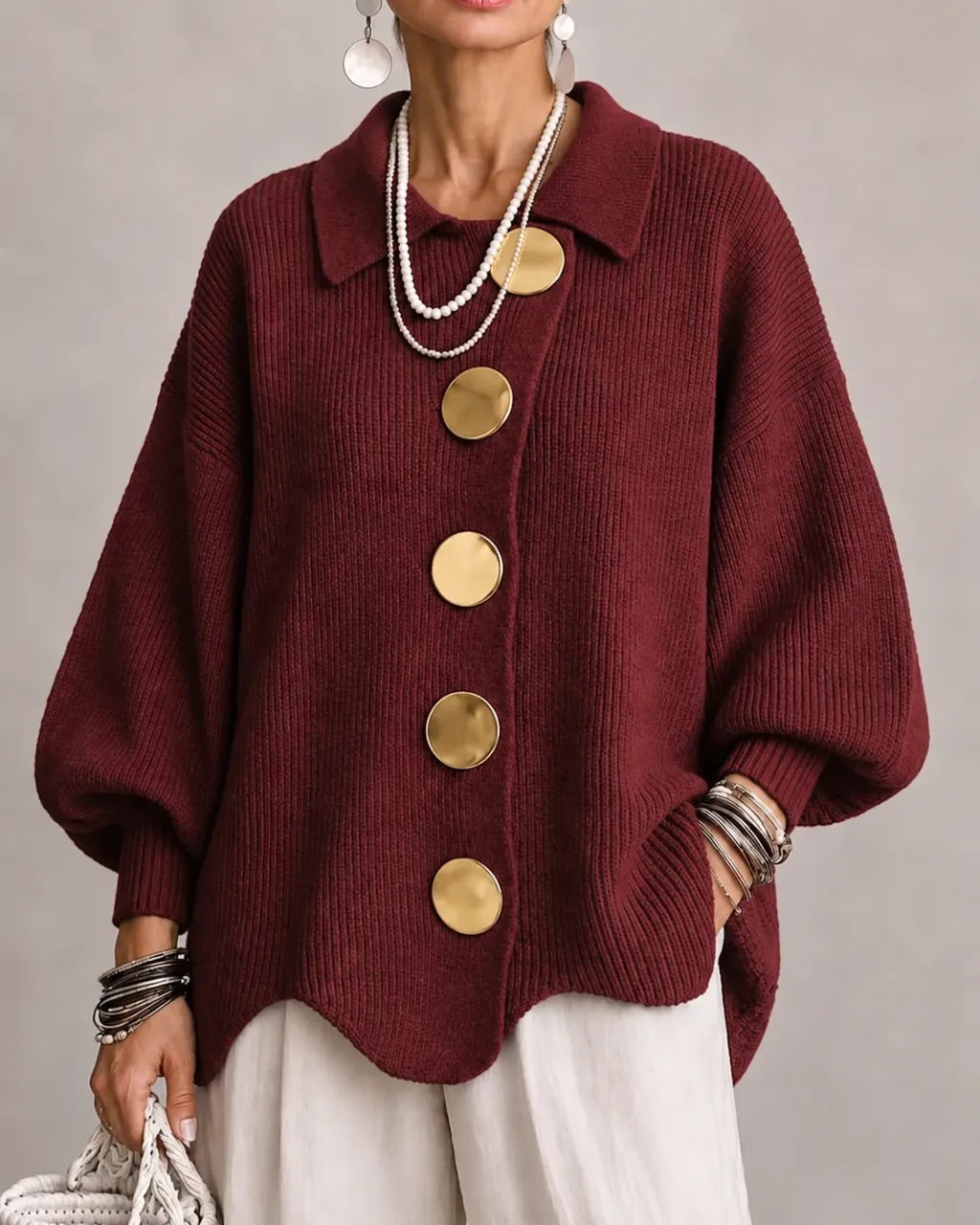 Burgundy ribbed cardigan on a model, with big gold buttons, pointed collar and scalloped hem, worn with white trousers. 19