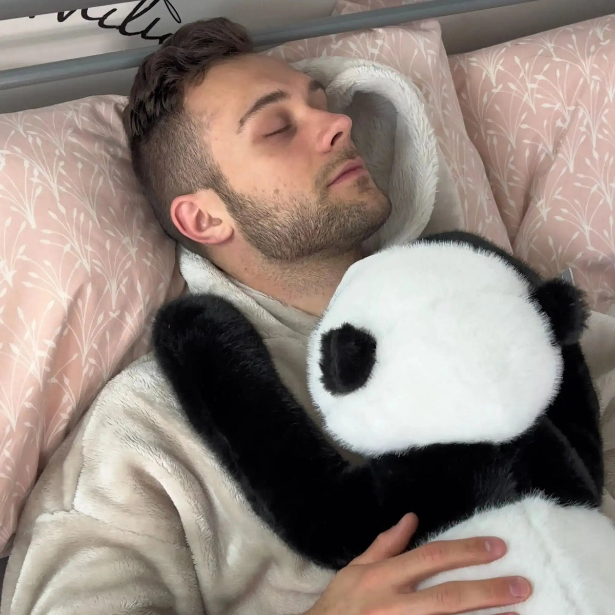 Man asleep in bed with pink patterned pillows, holding the panda plush against his chest with its arm over his shoulder. 11