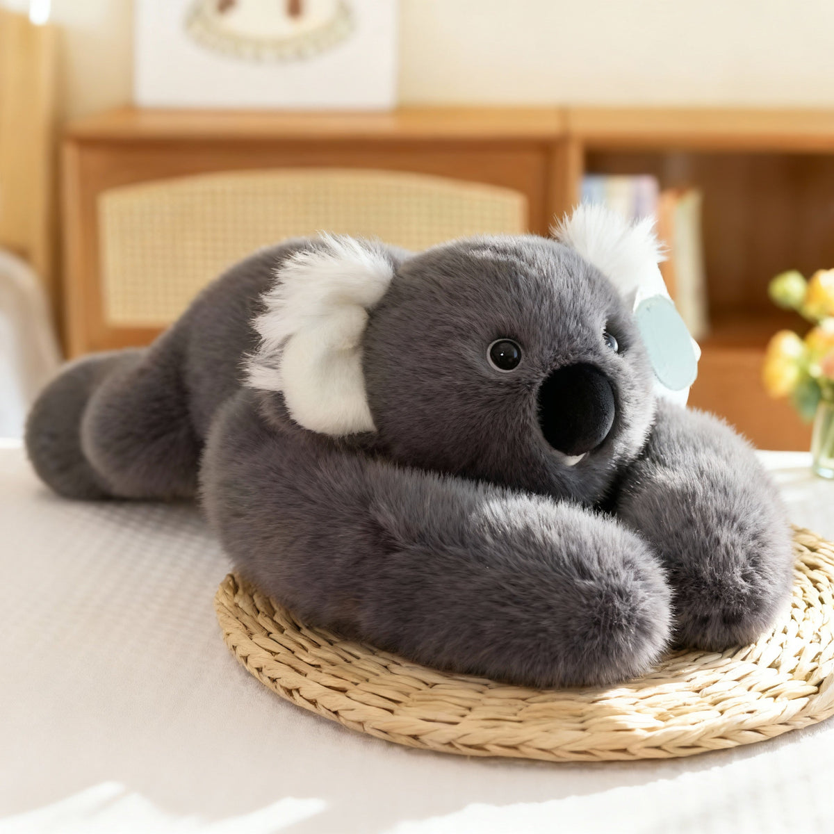 Grey koala plush with fluffy white ears and a large black nose, lying on a woven mat with long arms in front. 16