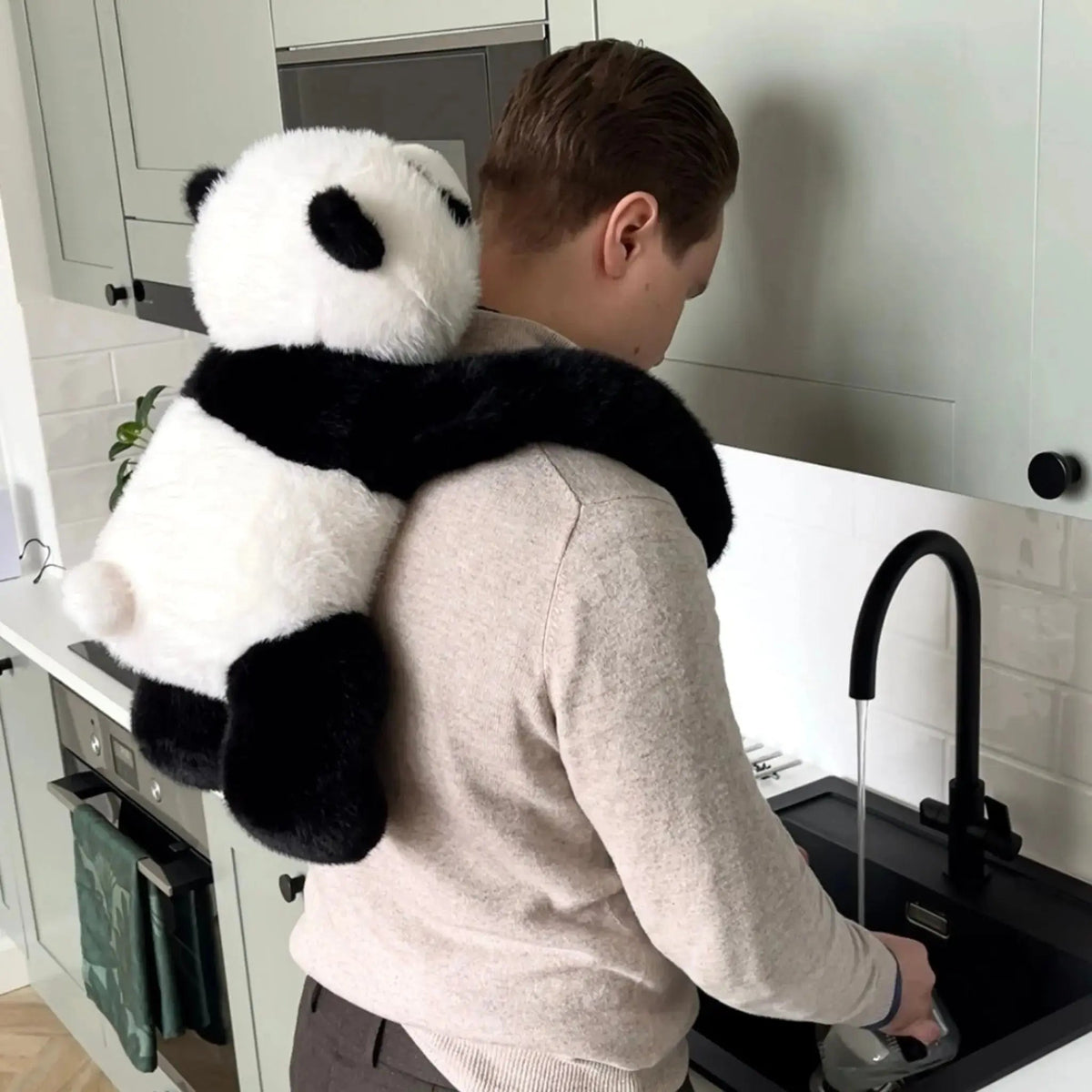 Man standing at a kitchen sink with the panda plush clinging to his back, long arms wrapped around his shoulders. 10