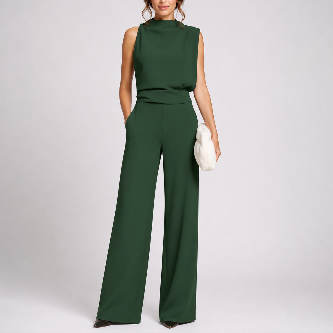 Model wearing the sleeveless jumpsuit in dark green with black pointed shoes, holding a fluffy cream clutch 1