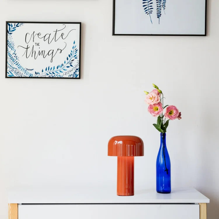 Orange lamp on a white sideboard beside a blue glass bottle of pink flowers, with framed prints above 20