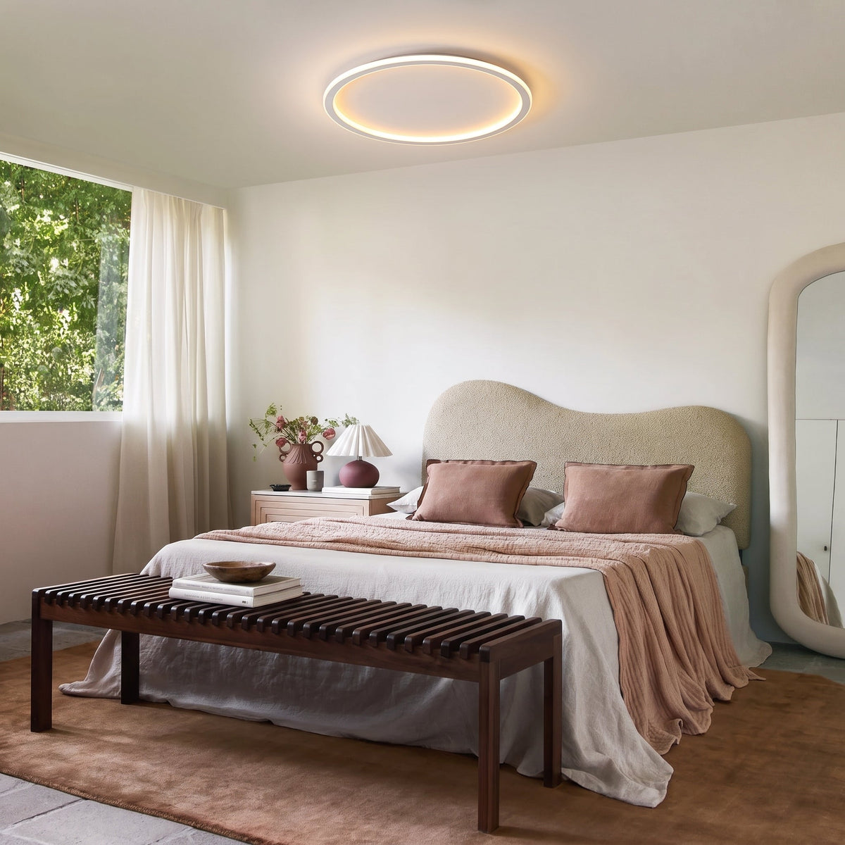 Ring ceiling light glowing above a bedroom with a curved boucle headboard, pink bedding, wooden bench and large floor mirror. 11