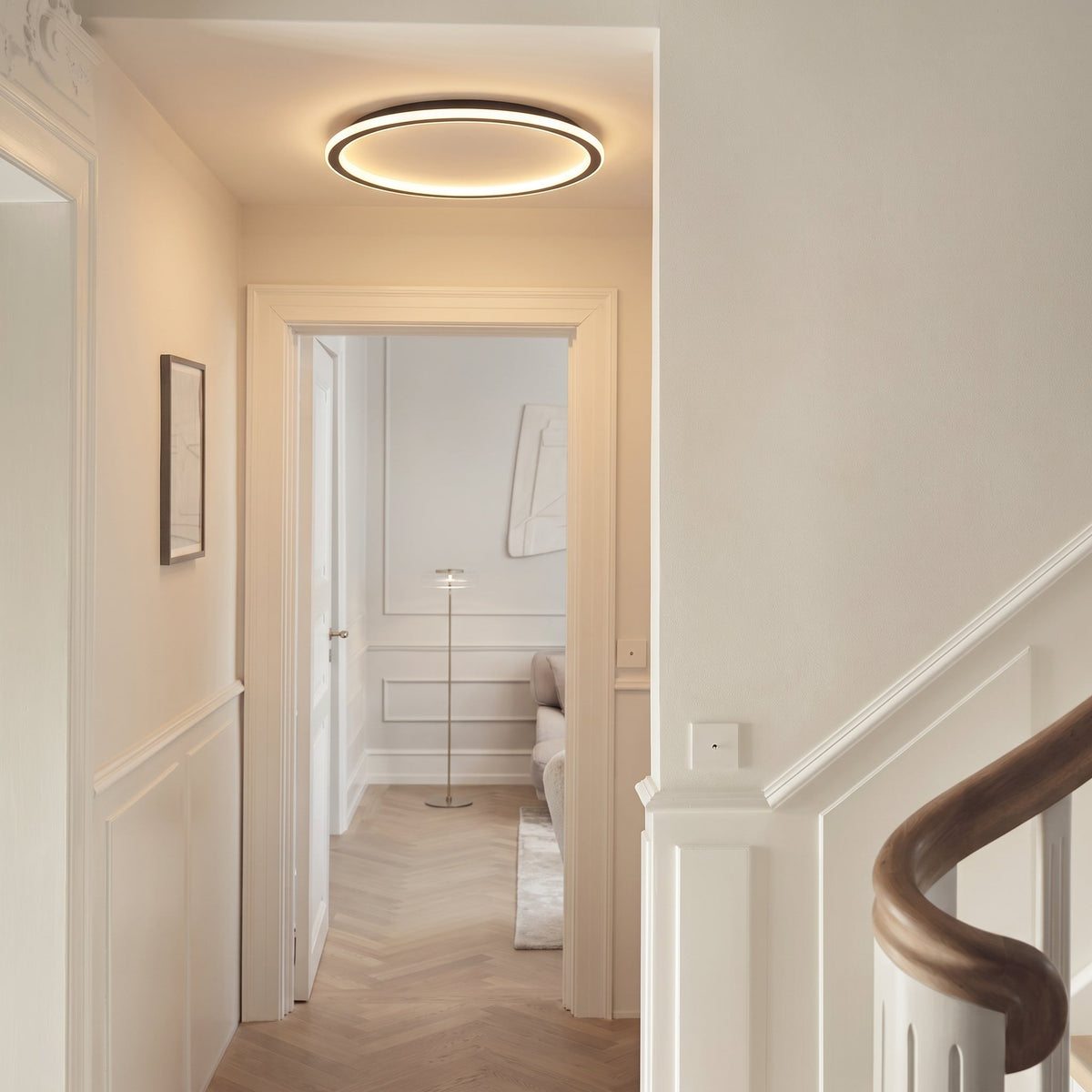 Lit ring light fitted in a hallway ceiling, with panelled walls, a staircase handrail and a doorway to another room. 5