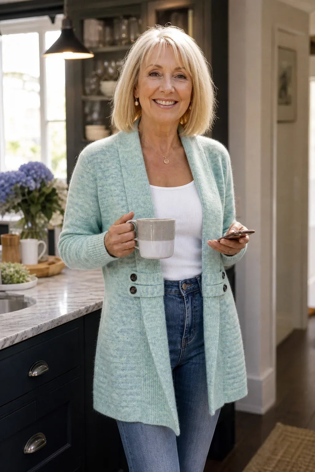 Older woman smiling in a kitchen, wearing the light mint cardigan with a mug and phone 4