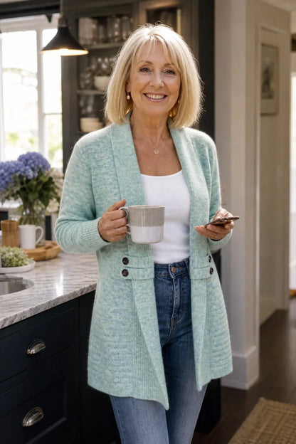 Older woman smiling in a kitchen, wearing the light mint cardigan with a mug and phone 4