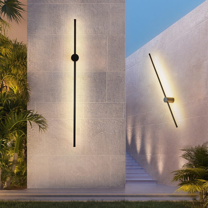 Slim black light bars on stone-clad walls, one upright and one tilted, glowing warmly beside garden steps and palm plants. 5