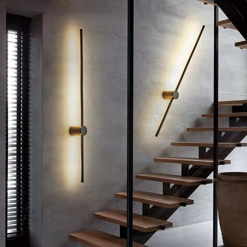 One vertical and one tilted black light bar with brass-coloured cylinder mounts on a grey concrete wall beside a wooden open staircase. 4