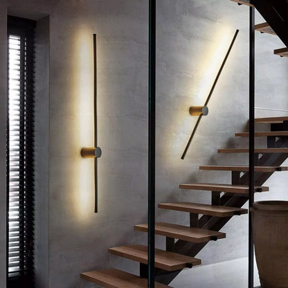 One vertical and one tilted black light bar with brass-coloured cylinder mounts on a grey concrete wall beside a wooden open staircase. 4