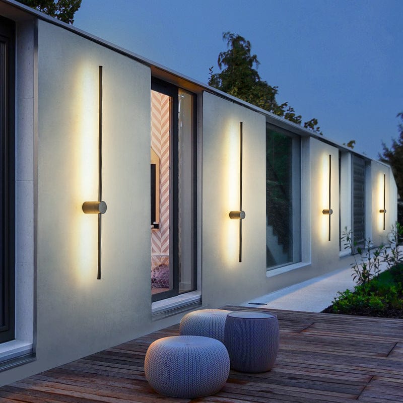 Four slim black light bars glowing warm white along a pale house wall at dusk, above a wooden deck with woven stools. 1