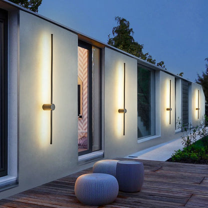 Four slim black light bars glowing warm white along a pale house wall at dusk, above a wooden deck with woven stools. 1