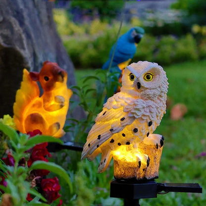 Glowing owl light in a flowerbed beside a lit squirrel light and a blue parrot ornament. 12