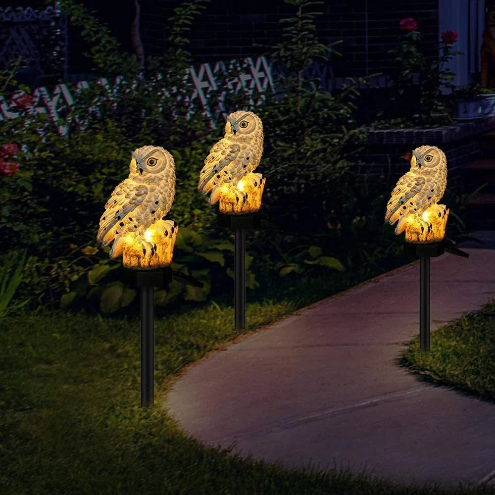 Three glowing owl solar lights on stakes lining a curved garden path at night. 3