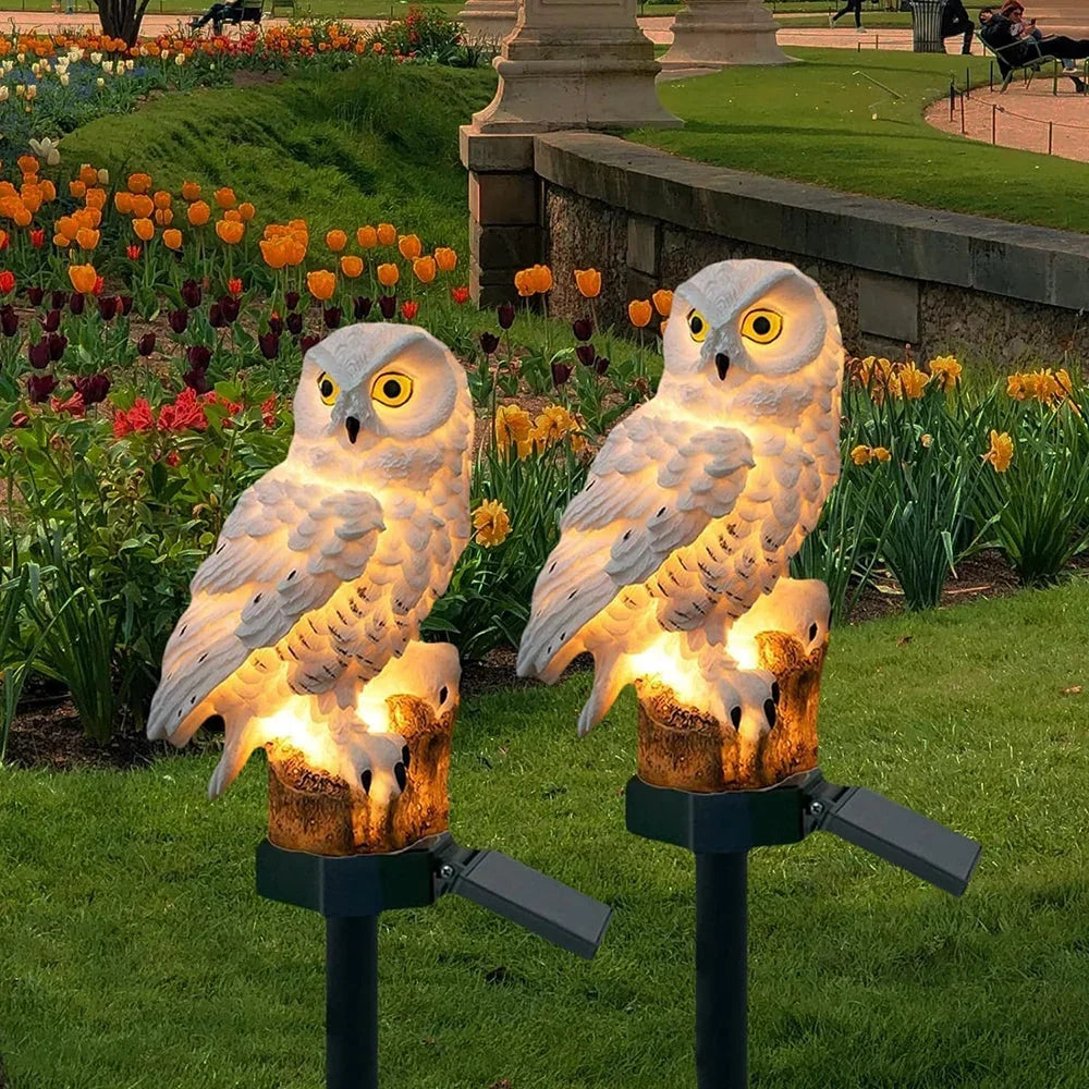 Two white owl solar lights glowing warmly on stakes in a flowerbed of orange tulips at dusk. 1