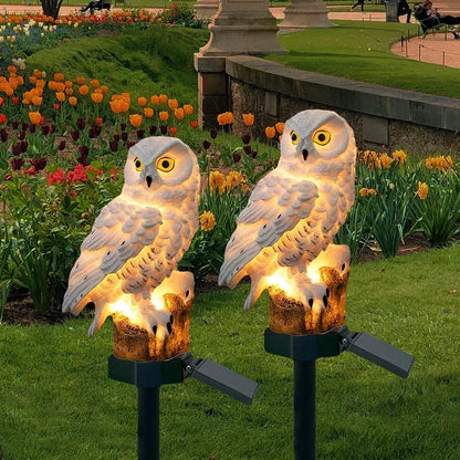 Two white owl solar lights glowing warmly on stakes in a flowerbed of orange tulips at dusk. 1