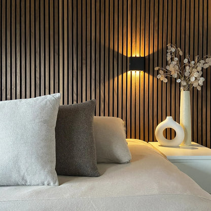 Black cube wall lamp glowing between slatted wooden wall panels above a bed with cushions and a bedside table with vases. 5