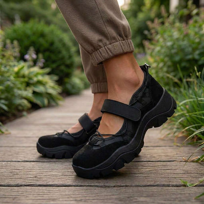 Black suede shoes with a strap and elastic laces, worn with khaki cuffed trousers on a wooden garden path. 10