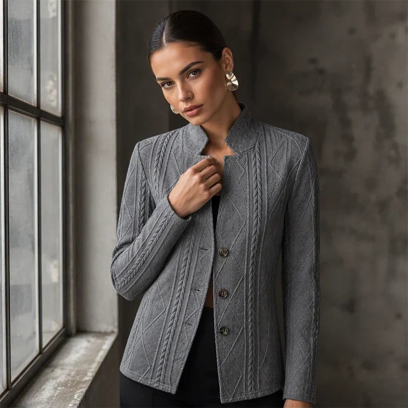 Woman in the grey textured blazer standing by a window, one hand on the stand collar, showing cable and diamond patterns. 10