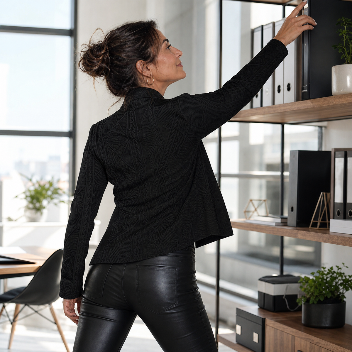 Back view of a woman in the black textured blazer and leather-look trousers, reaching up to a shelf of office files. 13