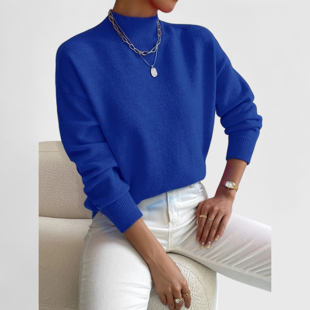 Model wearing the jumper in vivid royal blue with white jeans and silver layered necklaces, seated on a cream armchair. 11