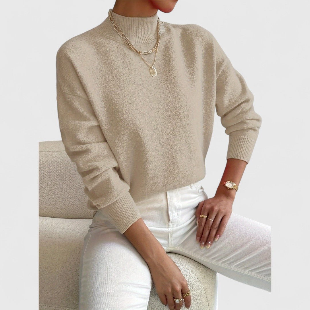 Model wearing the jumper in beige with white jeans and gold necklaces, seated against a cream bouclé chair. 7