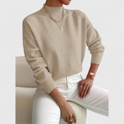 Model wearing the jumper in beige with white jeans and gold necklaces, seated against a cream bouclé chair. 7