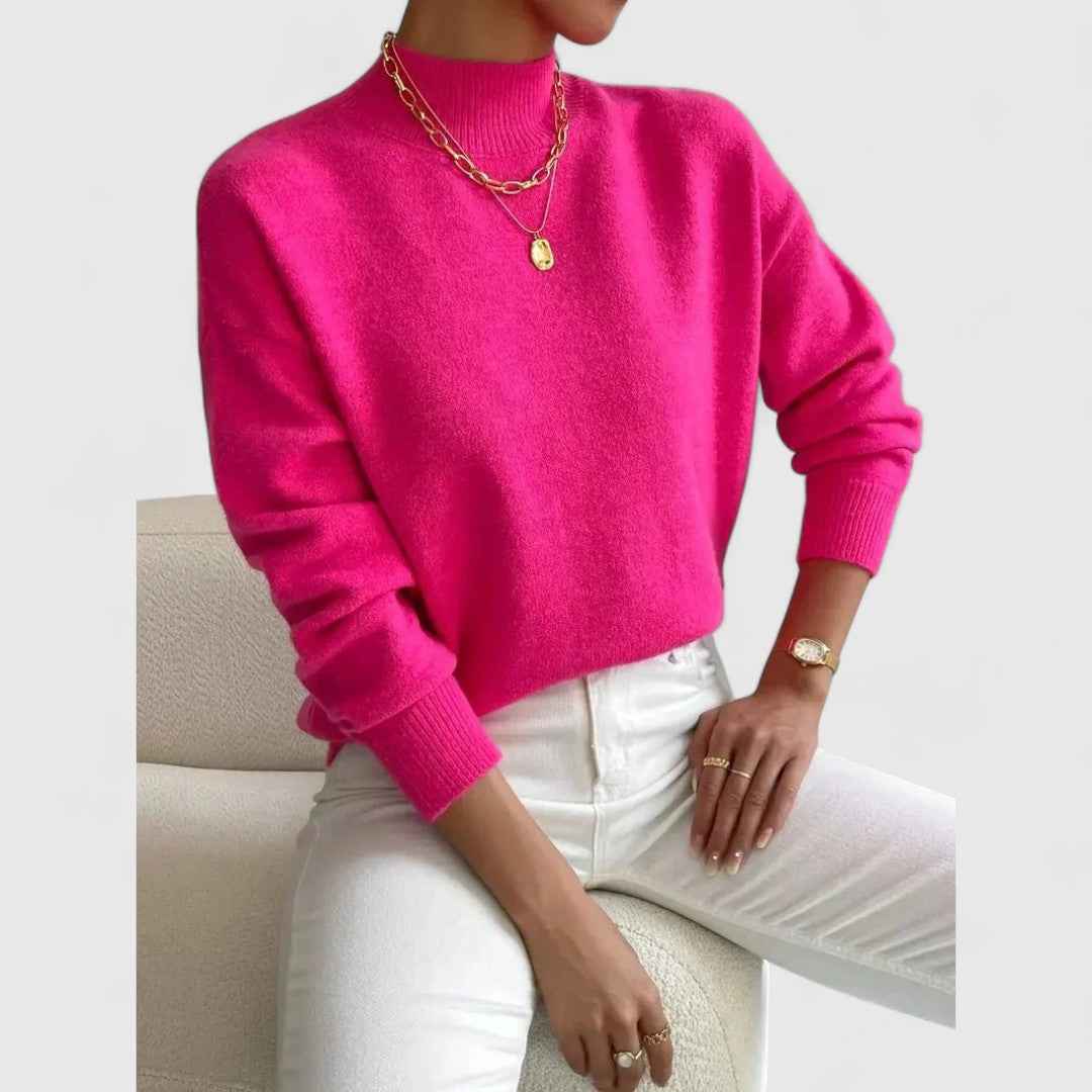 Model wearing the jumper in bright fuchsia pink with white jeans, gold necklaces and a watch, seated on a cream chair. 9