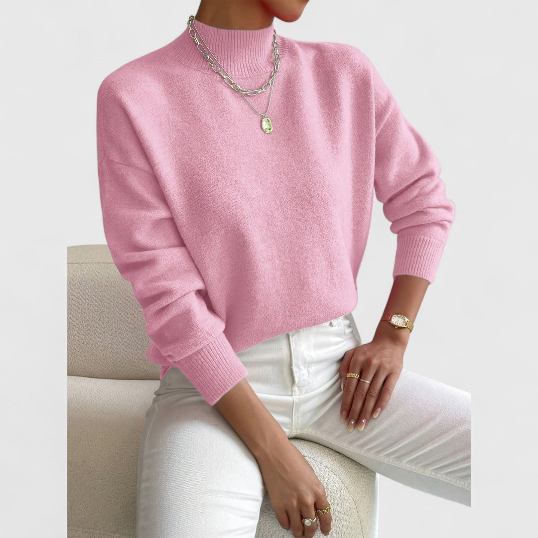 Model seated in a cream armchair wearing the jumper in light pink, with ribbed cuffs and a mock neck visible. 5