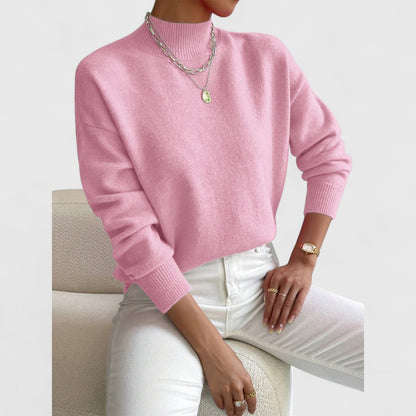 Model seated in a cream armchair wearing the jumper in light pink, with ribbed cuffs and a mock neck visible. 5