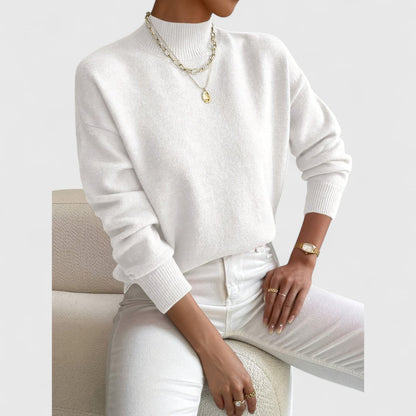 Model wearing the jumper in white with white jeans and gold layered necklaces, seated on a cream bouclé armchair. 4