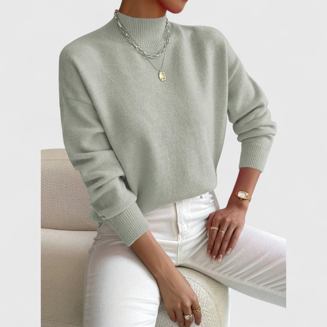 Slightly closer view of the sage green jumper worn by a seated model, showing the ribbed funnel neck and cuffs. 3