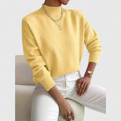 Model wearing the jumper in soft yellow with white jeans, one hand resting on her knee beside a cream chair. 6
