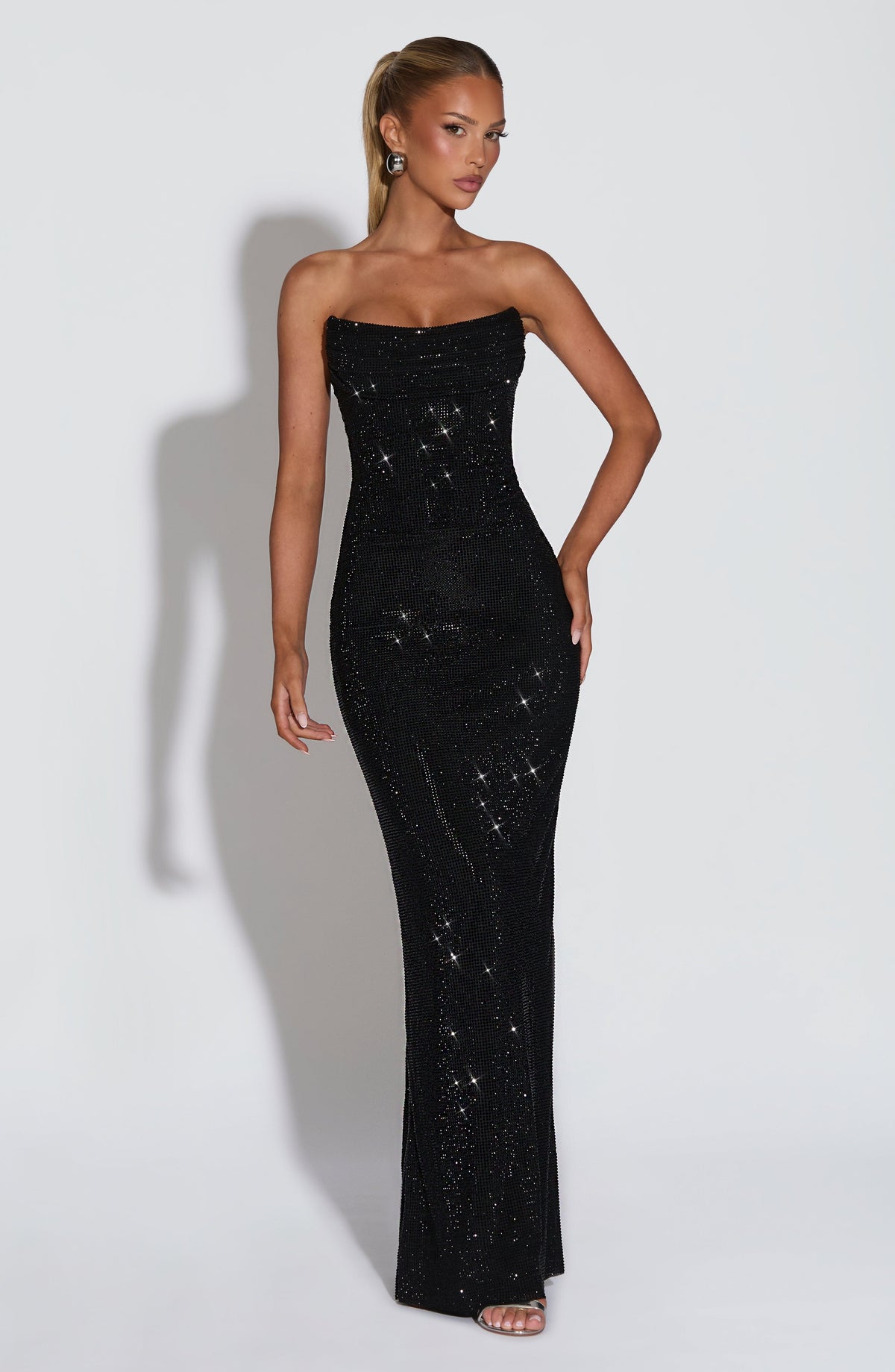 Model wearing the black strapless rhinestone maxi dress, full-length front view, hand on hip, with a draped neckline and fitted silhouette. 22