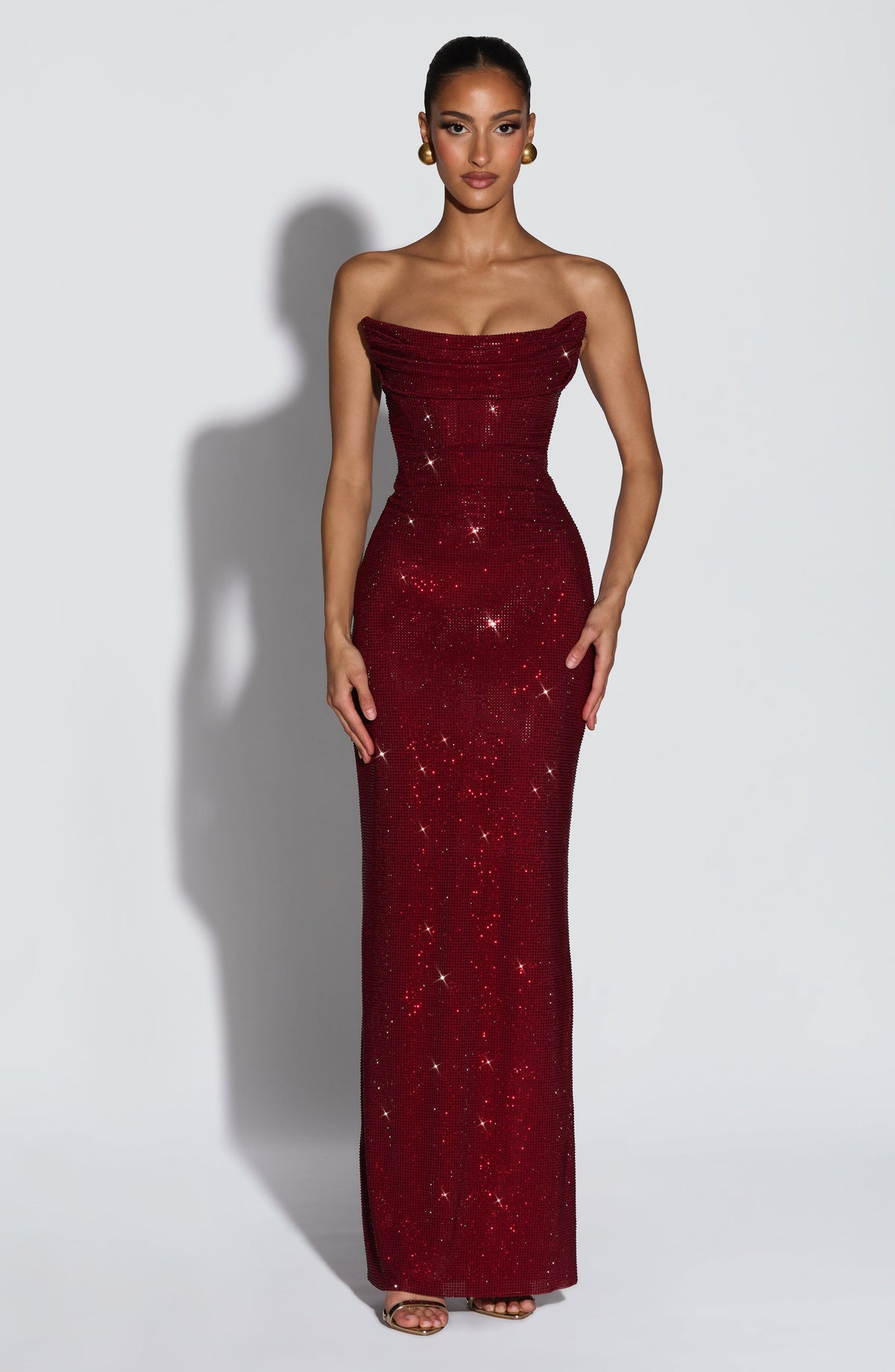 Model facing forward in the floor-length strapless burgundy rhinestone dress, with draped corset bodice and gold earrings. 1