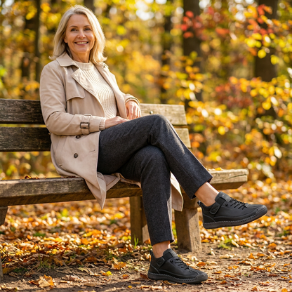 Woman in a beige trench coat and grey trousers seated on an autumn bench, wearing black lace-up trainers. 17