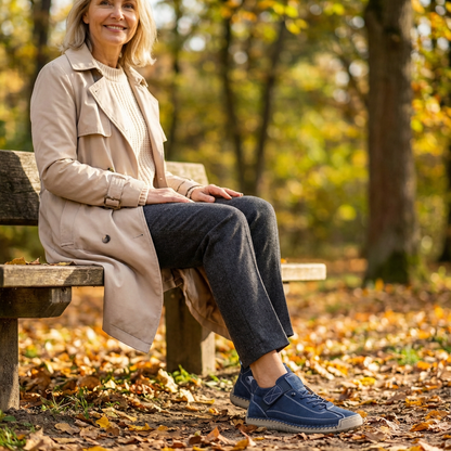 Smiling woman in a beige trench coat sits on an autumn park bench wearing navy blue suede-look walking trainers with a hook-and-loop strap. 21