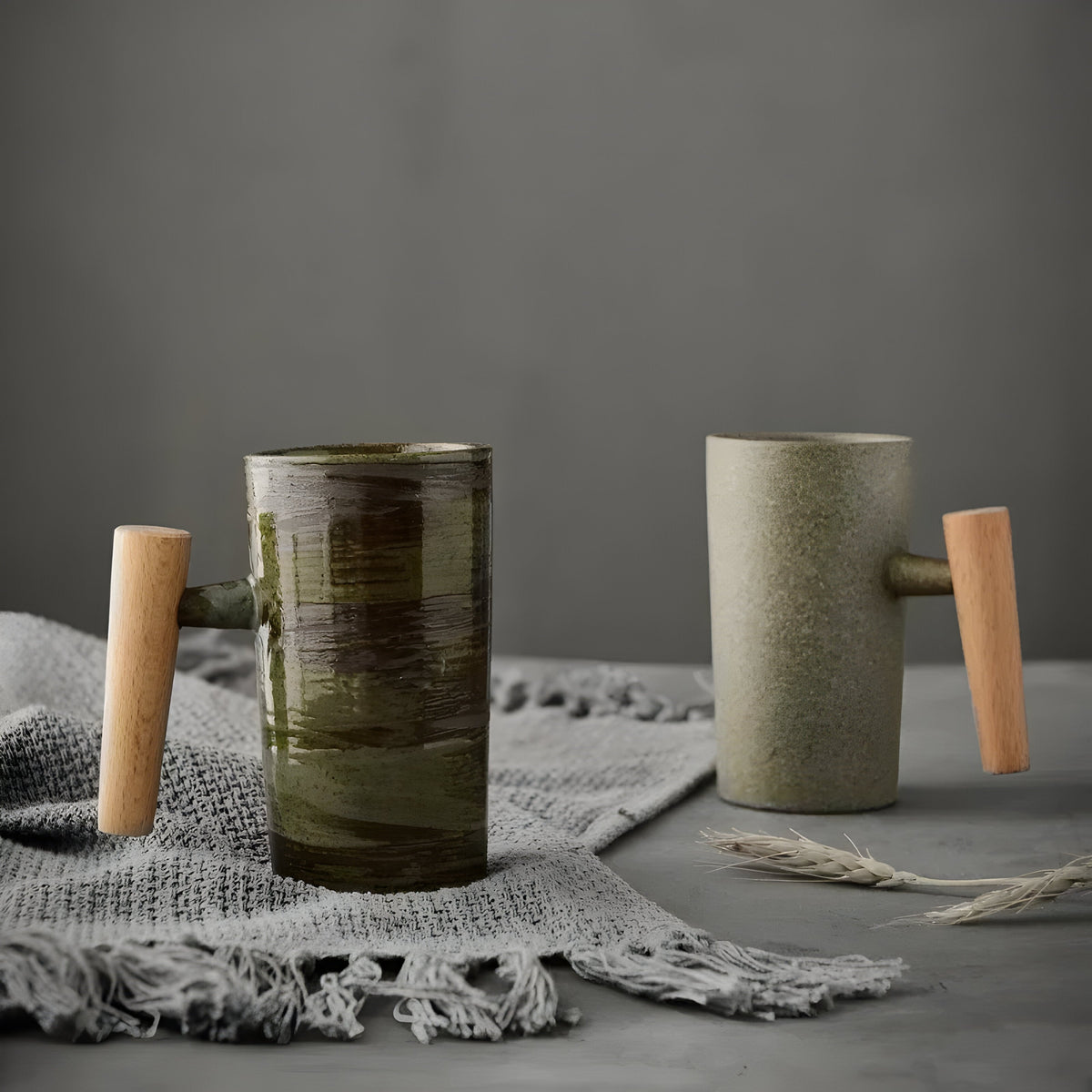 Two tall stoneware mugs with wooden handles, one brushed green-brown, one sandy beige, on a grey fringed cloth 1