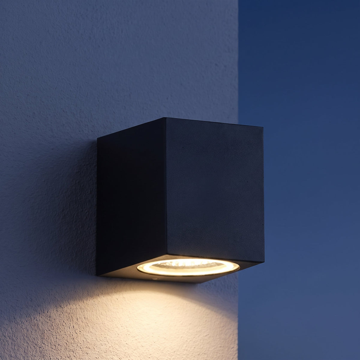 Black cube wall lamp casting a warm downward beam on a textured wall at dusk 2